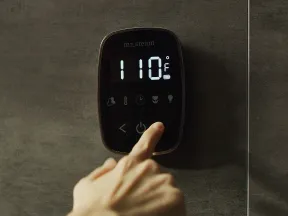 Hand pressing power button on digital steam shower thermostat displaying 110°F on dark tiled wall.
