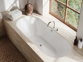 Modern bathroom with white built-in bathtub, natural stone surround, window, plants, and bath accessories.