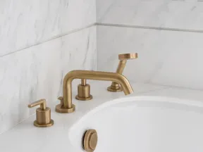 Modern bathtub with brushed gold faucet and handheld shower on white marble countertop and wall.