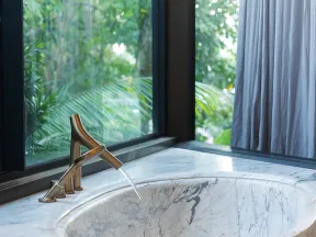 Modern marble bathroom sink with running water faucet next to large window overlooking greenery.