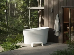 Modern white freestanding bathtub on wooden deck surrounded by forest greenery and attached sauna room.