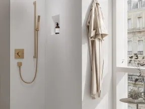 Minimalist bathroom shower with gold fixtures, beige robe, terrazzo floor, and large window overlooking city buildings.