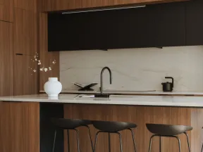 Modern kitchen with wood cabinets, marble countertop, black stools, and minimalist decor with white vase and flowers.
