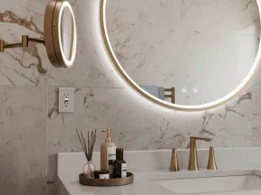 Modern bathroom with a round illuminated mirror, marble walls, white countertop, and gold faucet fixtures.