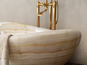 Close-up of a beige marble bathtub with a beige towel and vintage gold faucet on textured wall and wooden floor.