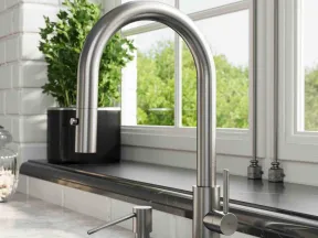 Modern stainless steel kitchen faucet and soap dispenser on a marble countertop by a window with greenery outside.