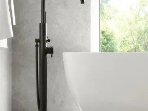 Modern black freestanding tub faucet next to a white bathtub in a minimalist bathroom with concrete walls and natural light.