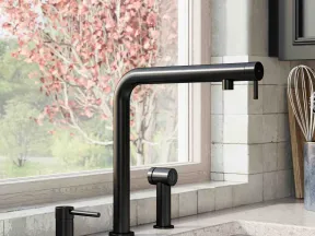 Modern black kitchen faucet with soap dispenser and sprayer by a window showing a blooming tree outside.