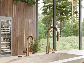 Modern kitchen with dual bronze faucets, stone countertop, wood cabinetry, and large windows overlooking greenery