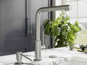 Modern stainless steel kitchen faucet and soap dispenser on white marble countertop with plant in background