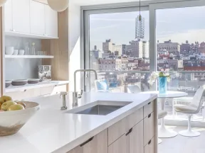 Bright modern kitchen with white countertops, wood cabinets, large window, city view, and dining area with chairs.