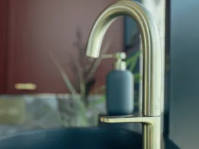 Modern brushed gold faucet over a black sink with a blurred background and soap dispenser.