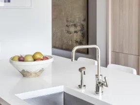 Modern kitchen countertop with stainless steel sink, chrome faucet, and a bowl of fruit on white surface.