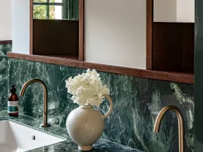 Modern bathroom with green marble countertop, dual brass faucets, white sinks, and a decorative white flower vase.
