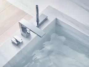 Close-up of modern chrome bathroom faucet and water flowing into a marble bathtub with light wooden flooring.