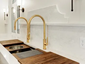 Modern kitchen sink area with dual gold faucets, marble countertops, wooden cutting boards, and wall sconces.