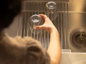 Person placing inverted glass on drying rack beside stainless steel kitchen sink under warm light.