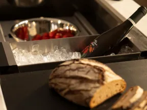 Loaf of crusty bread sliced on counter next to a wine bottle chilling in ice and bowls of fresh vegetables.