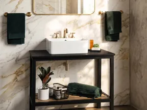 Modern bathroom with marble walls, black vanity, white sink, gold fixtures, and green towels with plant decor.