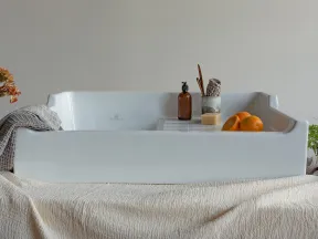 White ceramic bathtub tray with soap, brown bottle, brush, and oranges on a beige textured cloth.