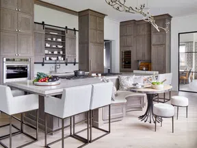 Modern kitchen with gray wooden cabinets, marble island, upholstered bar stools, and open shelf with dishes.