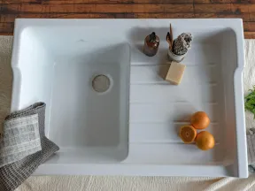 White ceramic kitchen sink with oranges, soap, and cloths on a wooden and fabric surface.