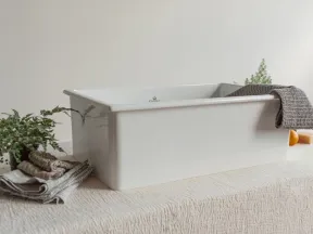 White ceramic farmhouse sink displayed on textured surface with towels, plant, soap, and gray cloth.