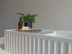 Modern white bathtub with textured sides, wooden tray holding soap, brown bottle, and a green potted fern plant.