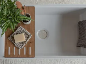 Minimalist white bathtub with wooden tray holding soap, toothbrush, and green plant, plus gray towel on tub edge.