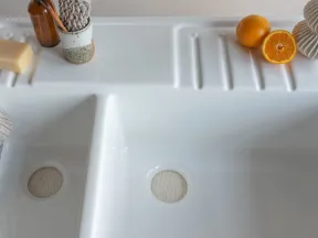 White double kitchen sink with a bar of soap, brown bottle, knitted cloth, and fresh oranges on the countertop.