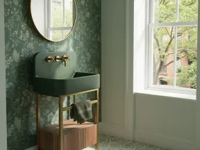 Green bathroom sink with gold fixtures and floral wallpaper next to a large arched window with natural light.