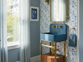 Modern blue sink with gold fixtures and wooden cabinet in a floral wallpaper bathroom with natural light.