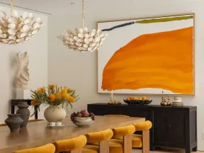 Modern dining room with mustard velvet chairs, wooden table, abstract orange wall art, and decorative lighting.