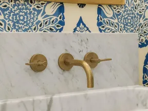 Marble sink with gold faucet set against blue and white floral wallpaper and a gold-framed mirror.