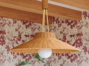 Woven rattan pendant lamp with white bulb hangs over green plant against floral wallpaper and wood beams.
