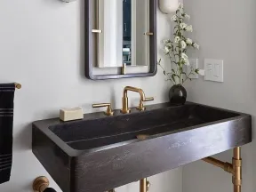 Modern bathroom with a black rectangular sink, brass faucet, wall sconces, mirror, and floral decor on a white wall.