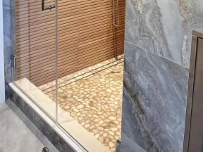 Modern shower with glass door, pebble stone floor, wooden slat wall, and gray marble tiles.