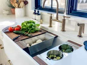 Modern kitchen sink with brass faucet, fresh vegetables on wooden cutting board, and stainless steel prep bowls.