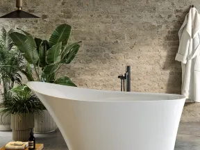Modern white freestanding bathtub in rustic bathroom with plants, wooden stool, and hanging robe.