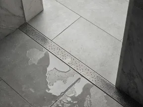 Modern bathroom floor with large gray tiles, wet area, and linear metal drainage grate beside marble walls.