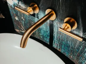 Modern bathroom faucet with brushed gold handles mounted on a dark textured wall above a white sink.