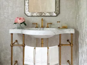 Elegant bathroom vanity with marble sink, gold fixtures, floral arrangement, mirror, and textured wall lighting.