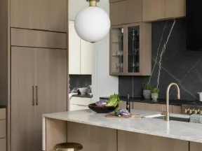 Modern kitchen with light wood cabinets, gold bar stools, marble island, black backsplash, and pendant light