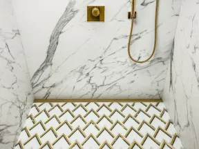 Elegant shower with white marble walls and geometric patterned floor tiles featuring gold accents and fixtures.