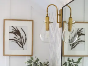 Modern bathroom counter with marble top, potted green plant, candle, brass wall light, and framed botanical print.