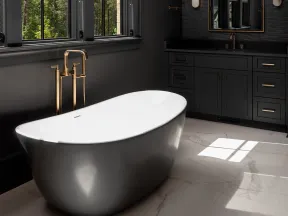 Modern bathroom with a sleek gray freestanding tub, brass fixtures, dark cabinetry, and marble flooring.