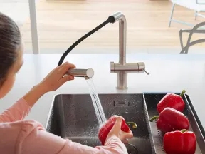 Person washing red bell peppers under a modern kitchen faucet over a black sink with natural light.