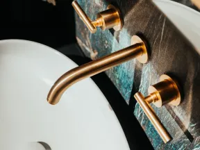 Modern bathroom sink with white basin and gold faucet handles mounted on dark textured wall.