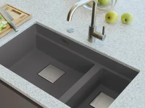 Modern double-basin gray kitchen sink with stainless steel faucet on light granite countertop with green apples and cutting board.