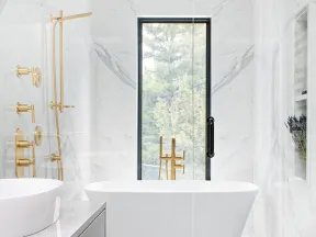 Modern white marble bathroom with freestanding tub, gold fixtures, large window, and vessel sink on gray vanity.